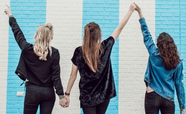 three women holding hands and putting them in the air in front of a brick wall with blue and white stripes